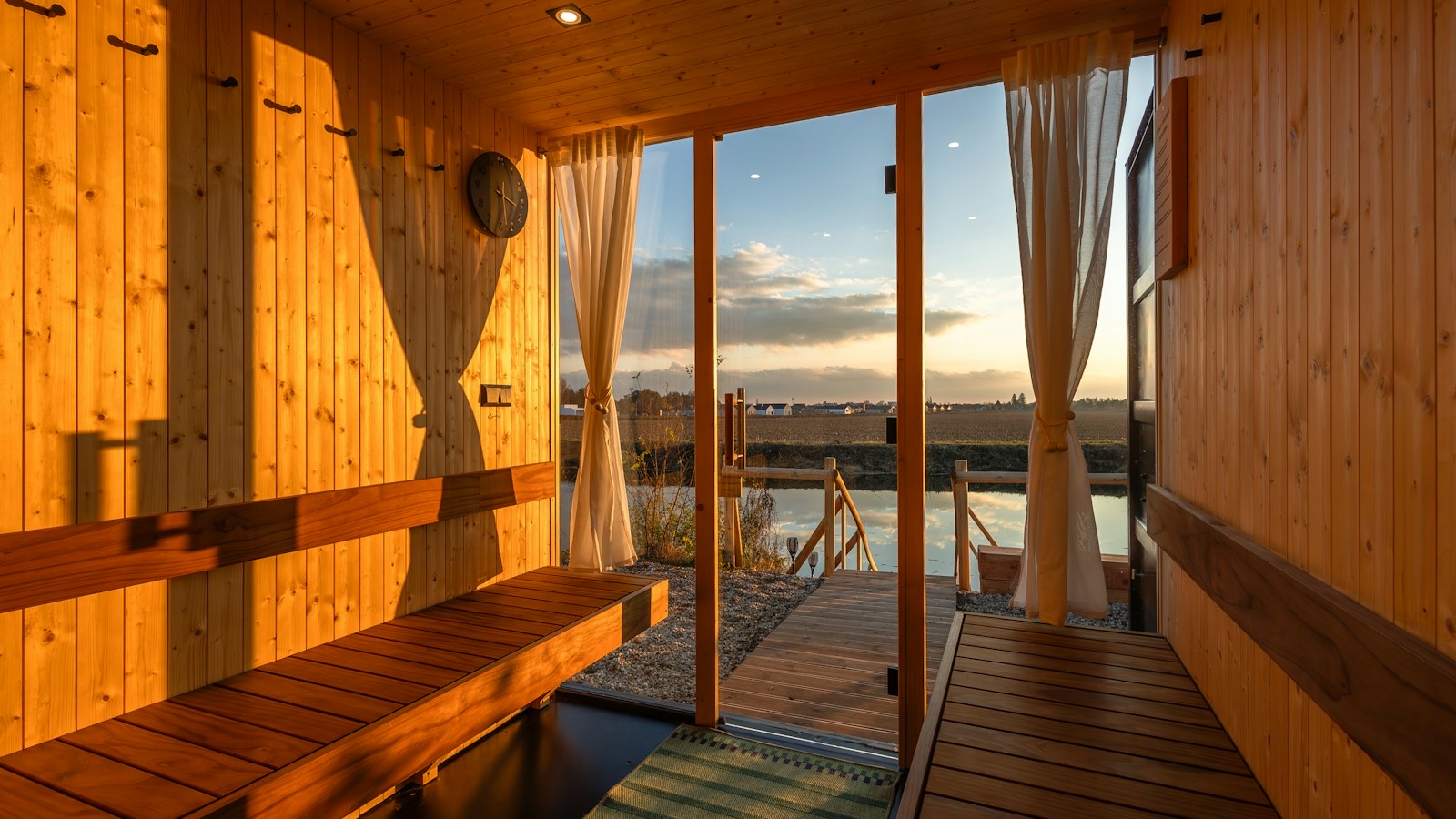 Houten saunaruimte met banken en een open deur naar een steiger aan het water bij zonsondergang